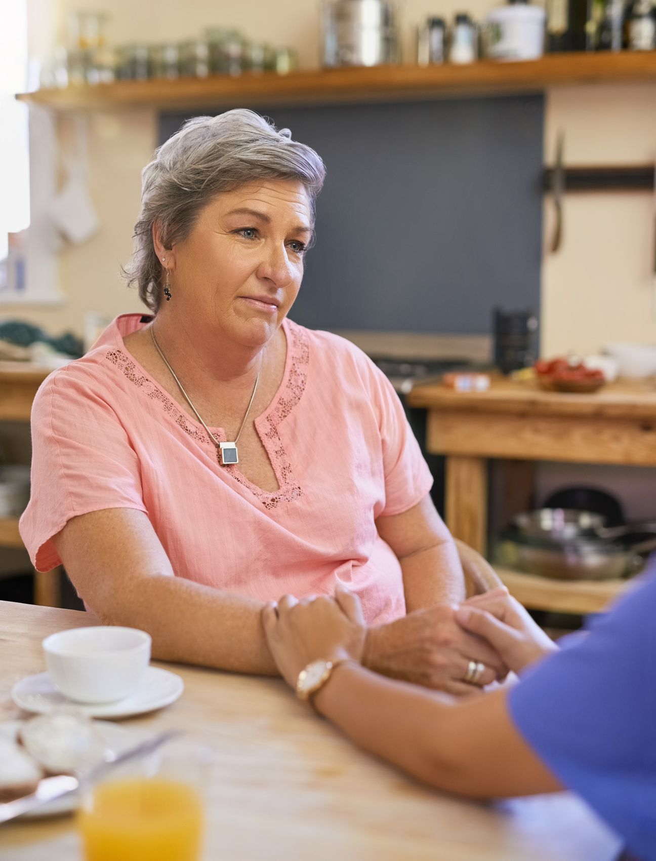 Older woman at her kitchen table with a caregiver's hand resting on hers